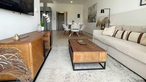 Uma sala de estar moderna com uma mesa de centro de madeira, um sofá bege e uma área de refeições ao fundo.