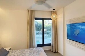 A hotel room with a bed, a television, a ceiling fan, and a view of the outdoors through a large window.