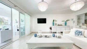 A modern living room with a white sofa, a coffee table, and decorative items. There is a TV mounted on the wall and a balcony visible through the glass doors.
