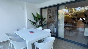 Une pièce moderne avec une table et des chaises blanches, une grande fenêtre donnant sur l’extérieur et une porte-fenêtre coulissante qui mène à un balcon.