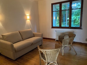 Sala de estar com sofá bege, cadeiras de vime, radiador e janela com vista para vegetação.
