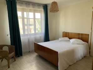 Quarto com cama de madeira, roupa de cama branca, uma cadeira de vime e uma janela com cortinas.
