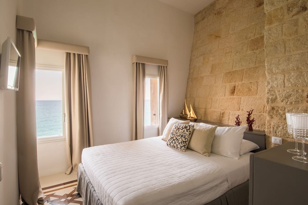 A hotel room with a large bed, a stone wall, and a view of the sea.