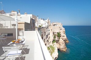 A seaside terrace with dining tables and chairs, overlooking a rocky coastline and the sea.