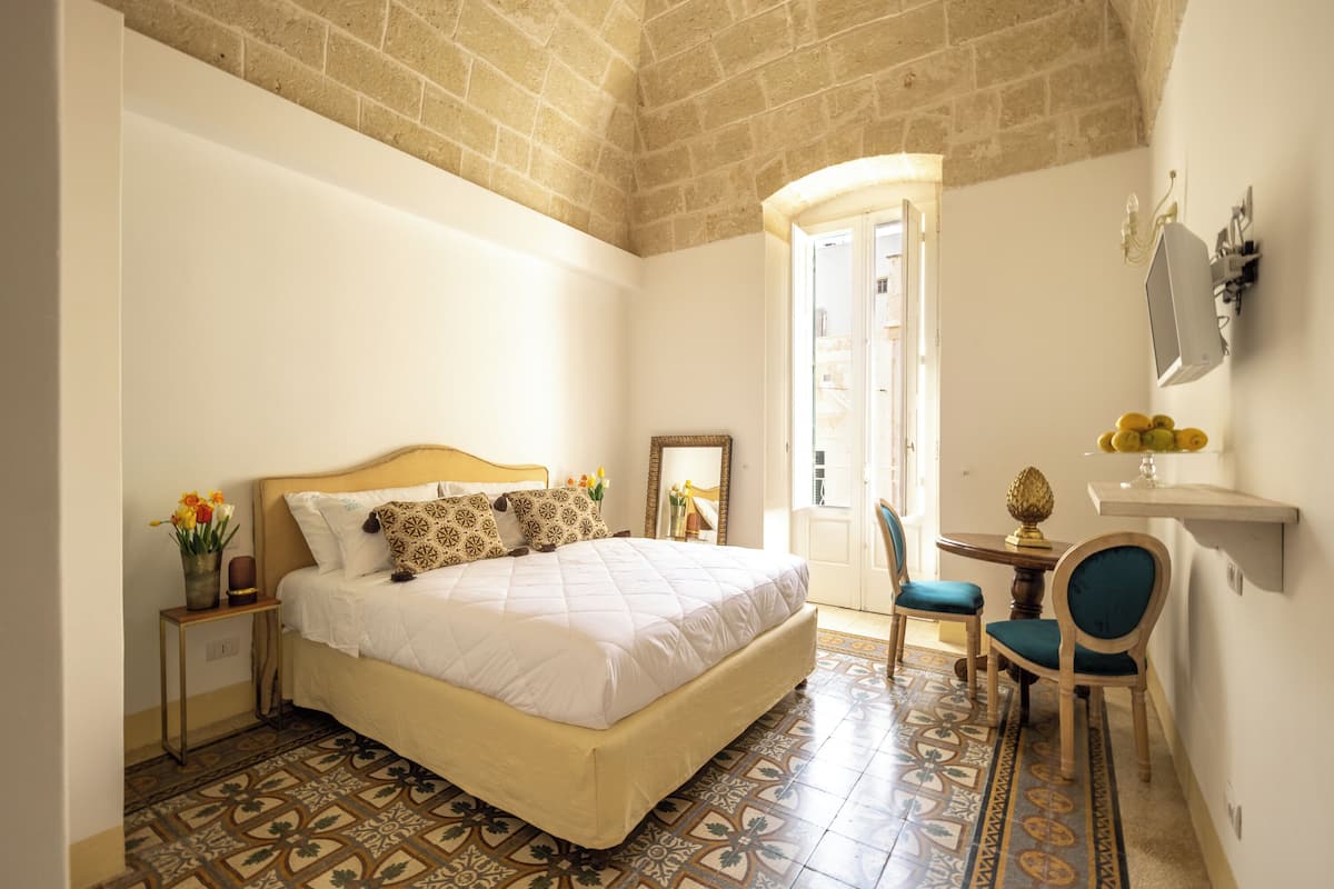 A bedroom with a large bed, a small table with chairs, a mirror, and a decorative vase with flowers.