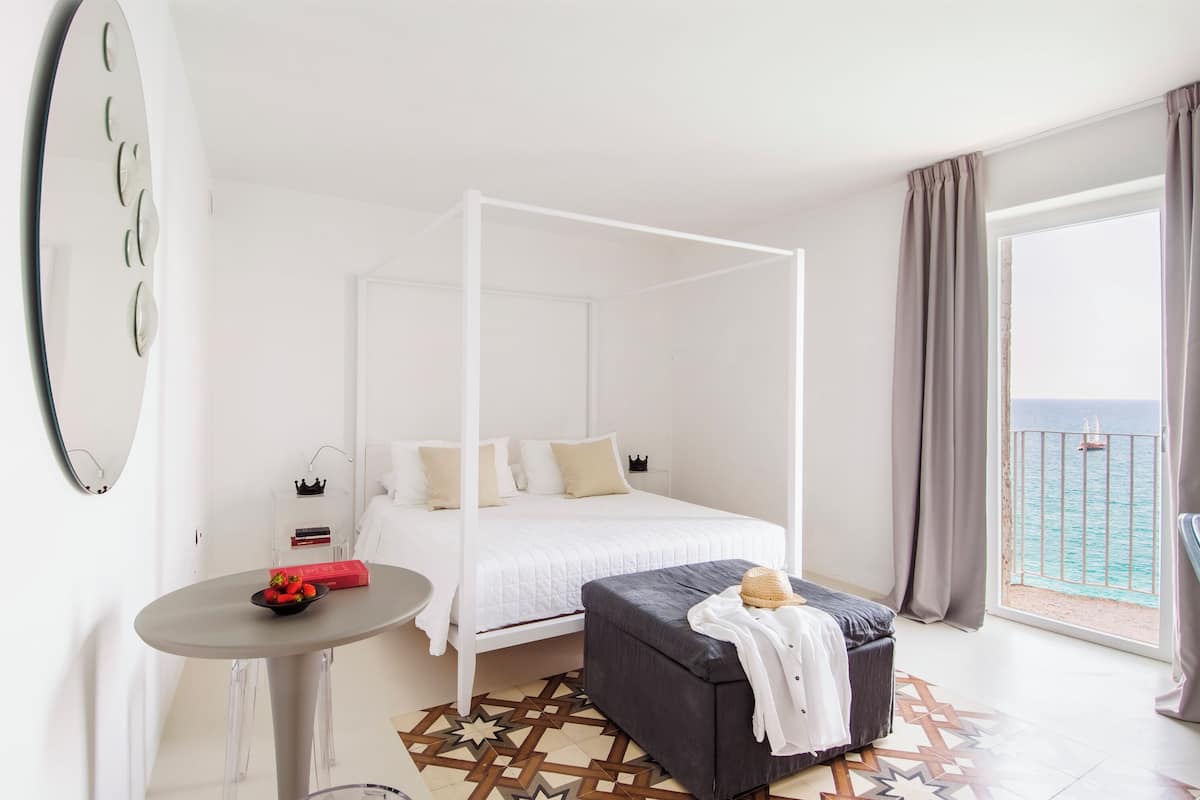 A modern bedroom with a four-poster bed, a small round table with a basket of fruit, and a view of the sea through large windows.