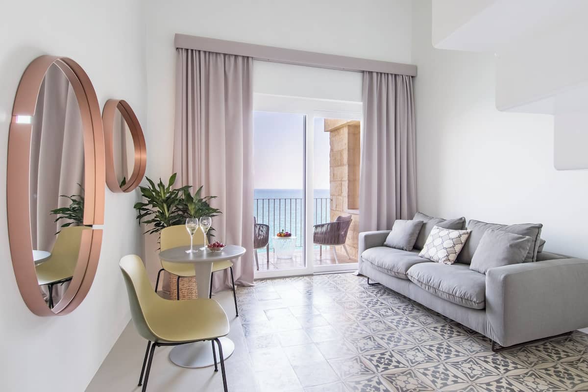 A modern living room with a grey sofa, a round table, and chairs, a balcony with curtains, and a large mirror on the wall.