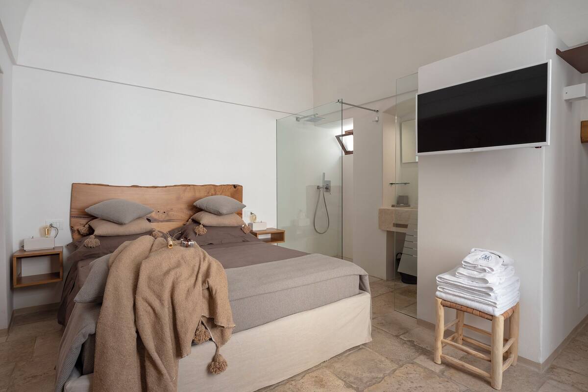 A modern bedroom with a large bed, a wooden headboard, a glass shower enclosure, a TV mounted on the wall, and a small wooden stool with folded towels.