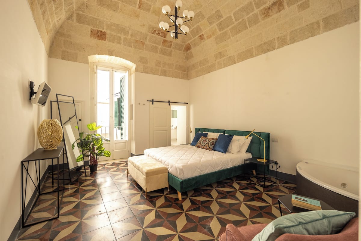 A bedroom with a geometric patterned floor, a bed with a green headboard, a bedside table, a lamp, a potted plant, and a view of the outside through a window.
