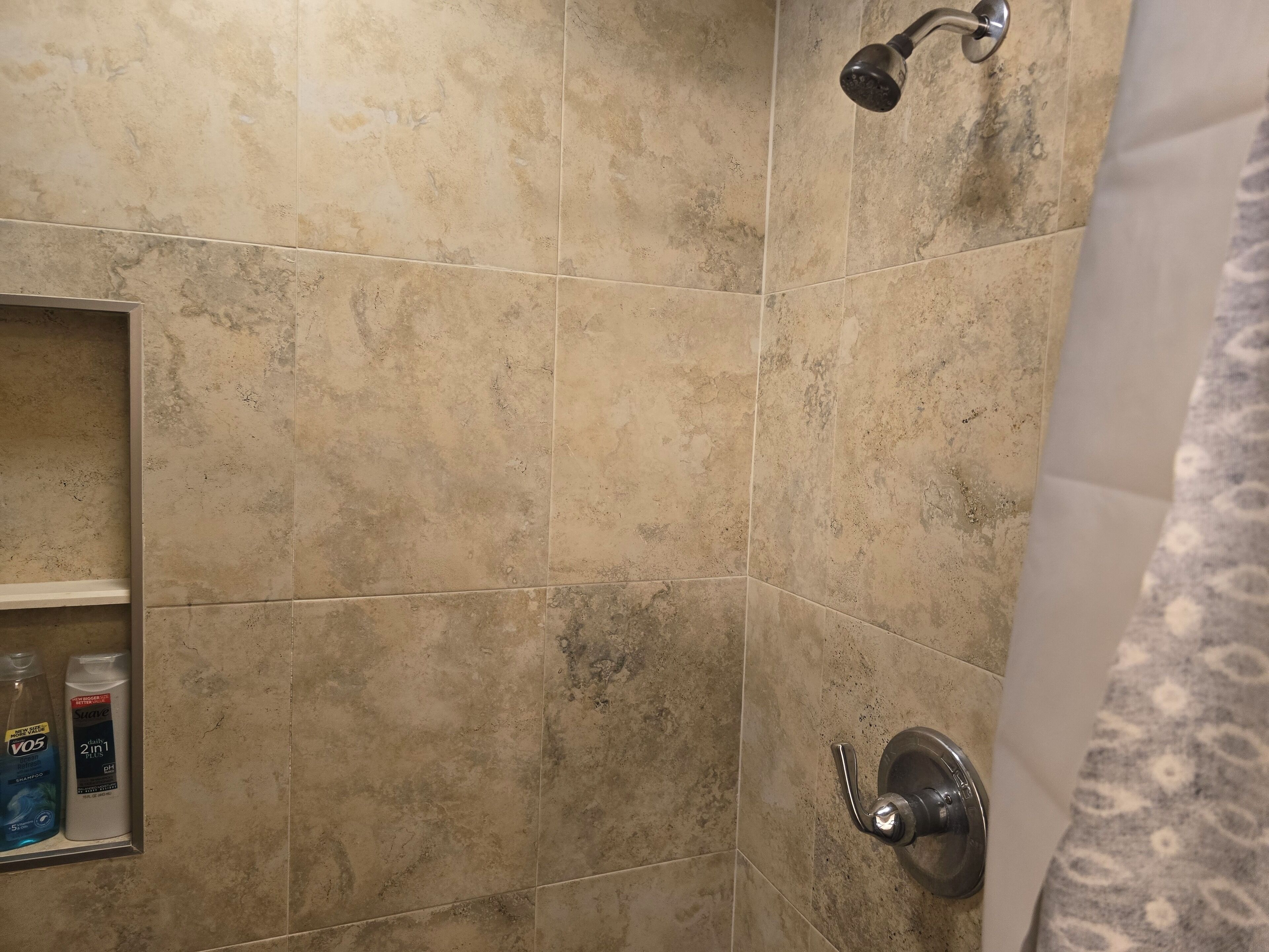 A bathroom with beige tiles, a showerhead, and a shelf with toiletries.