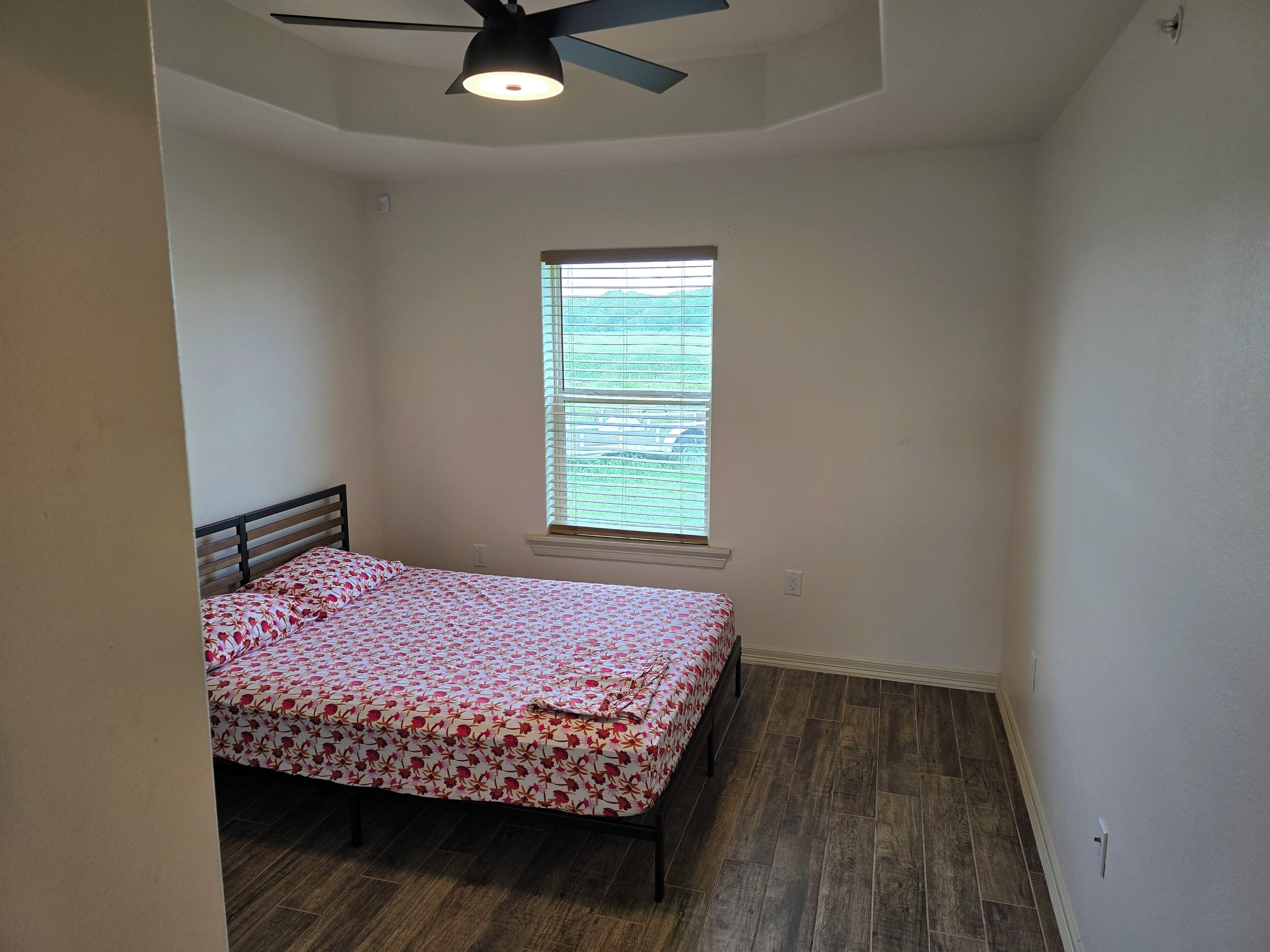 A bedroom with a bed, a window with blinds, a ceiling fan, and wooden flooring.