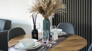 Una mesa de comedor preparada con platos y tazones blancos, cubiertos y un jarrón con flores secas.