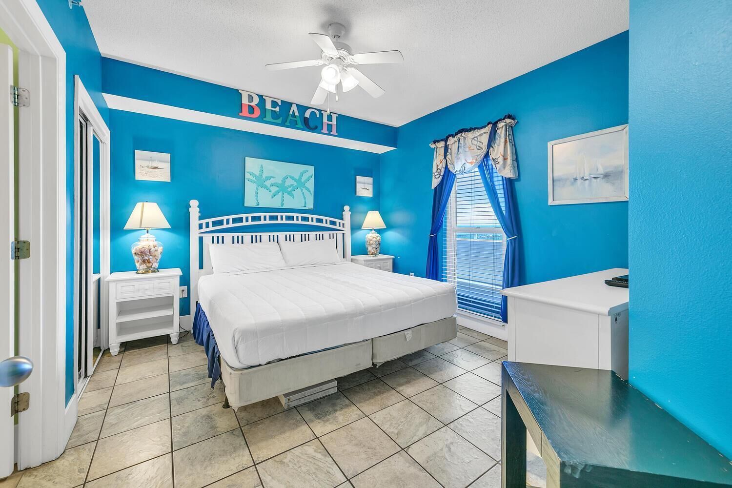A bedroom with a white bed, a ceiling fan, a blue wall with the word "BEACH" above it, and a window with blue curtains.