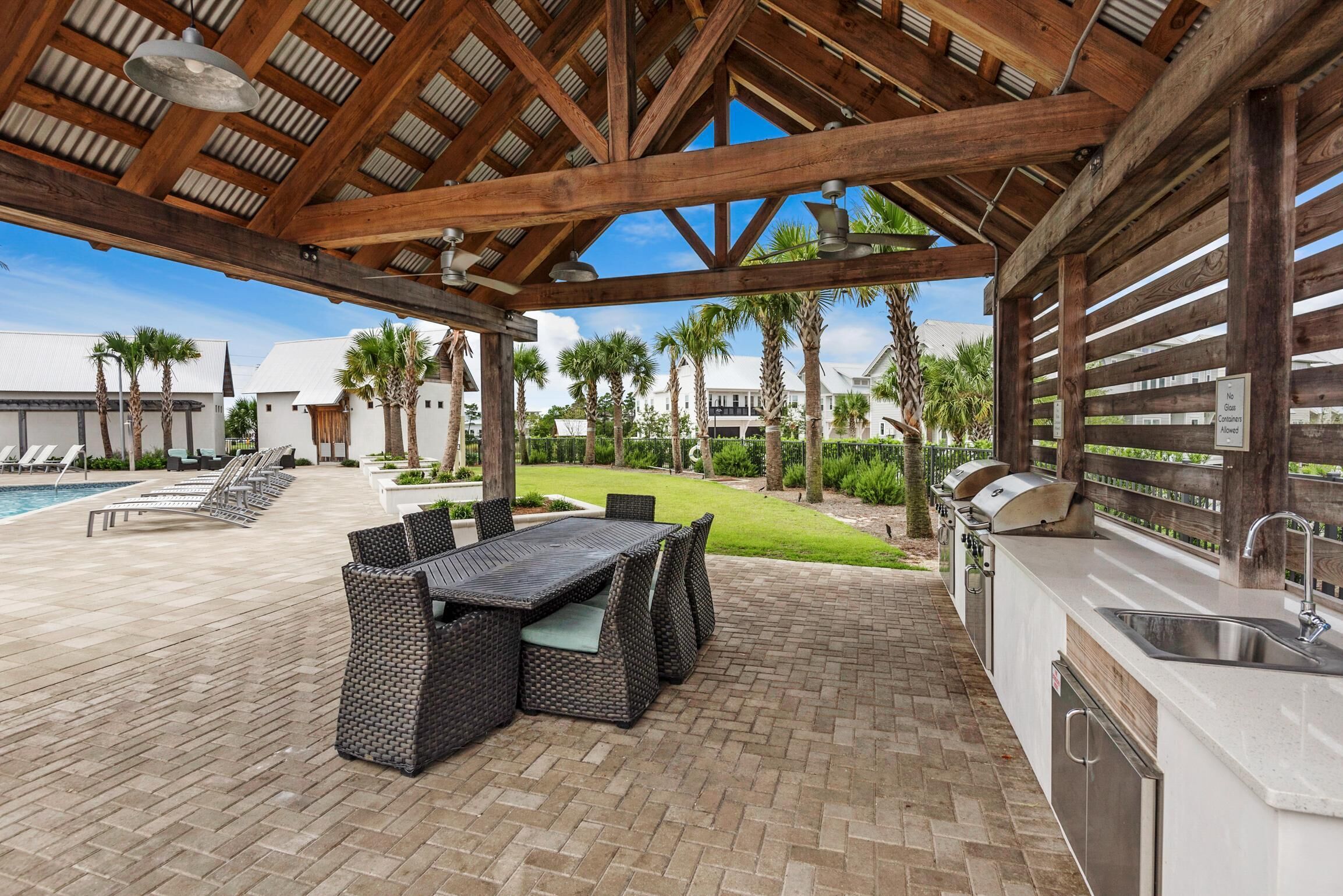 An outdoor patio with a brick floor, a dining area with wicker furniture, a grill station, and a pool in the background.