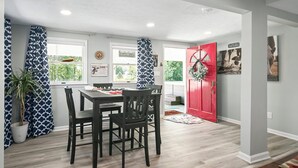 A dining area with a wooden table and chairs, a red door, and a window with a view of greenery.