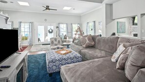 A spacious living room with a grey couch, a patterned ottoman, a flat-screen TV, and a ceiling fan.