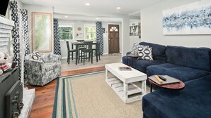 A living room with a blue sofa, a white coffee table, a patterned armchair, and a dining area with a wooden table and chairs.