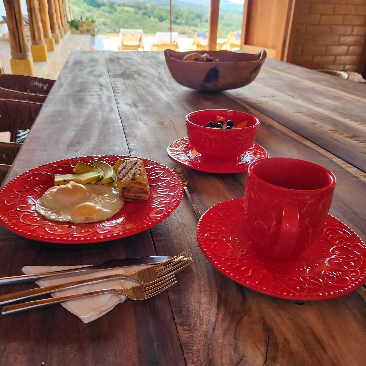 A wooden table with a red plate of food, a red cup, and a bowl of fruit.
