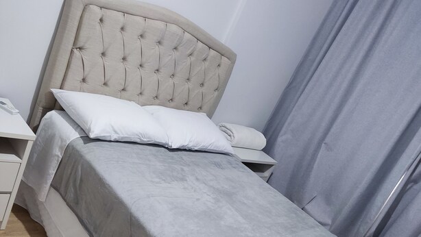 A neatly made bed with a grey bedspread and two pillows, a bedside table with a lamp, and a grey curtain. - Apartamento Completo Mobiliado com ar Condicionado Quente Frio (Country Cascavel)