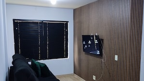 A modern living room with a black sofa, a wooden accent wall, a mounted TV, and a window with blinds. - Apartamento Completo Mobiliado com ar Condicionado Quente Frio (Country Cascavel)