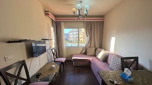 Ein Wohnzimmer mit einer Couch, einem an der Wand montierten Fernseher, einem Tisch mit einem Laptop und einem Fenster mit Vorhängen.