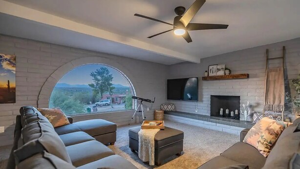 A modern living room with a large arched window, a fireplace, a ceiling fan, and a flat-screen TV. - Hilltop Private Resort Home with SPARKLING POOL (Tucson)