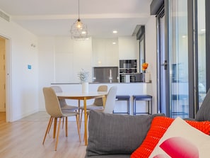 A modern kitchen with white cabinets, a dining area with a white table and chairs, and a sofa with a red and white cushion.