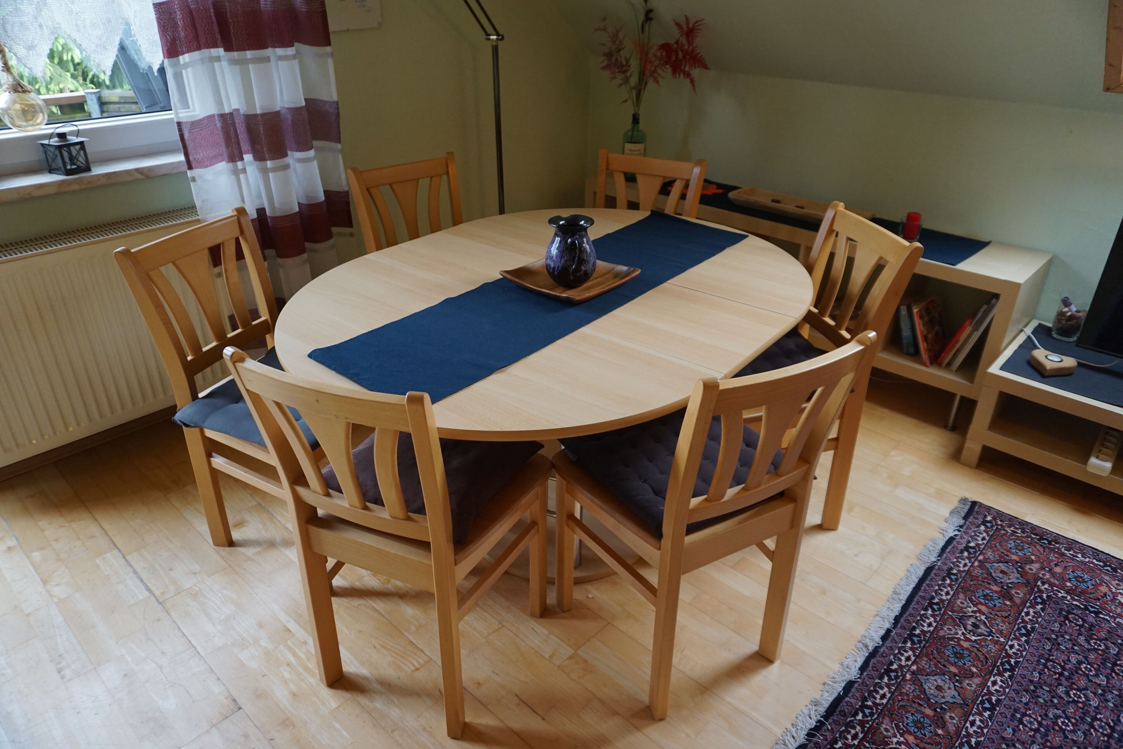 A round wooden dining table with blue placemats, surrounded by wooden chairs with cushions.