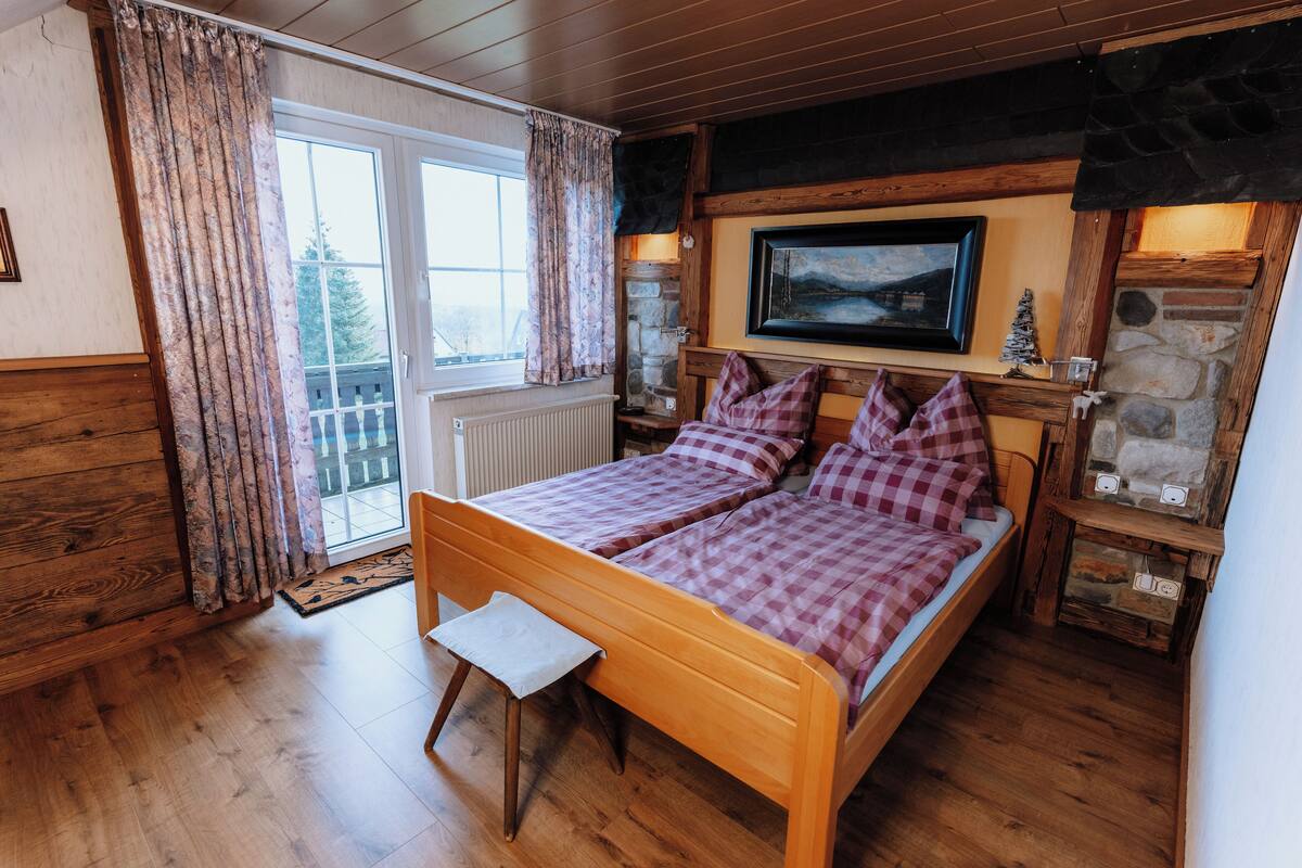 A cozy bedroom with a wooden bed, a small stool, a stone fireplace, and a large window with curtains.