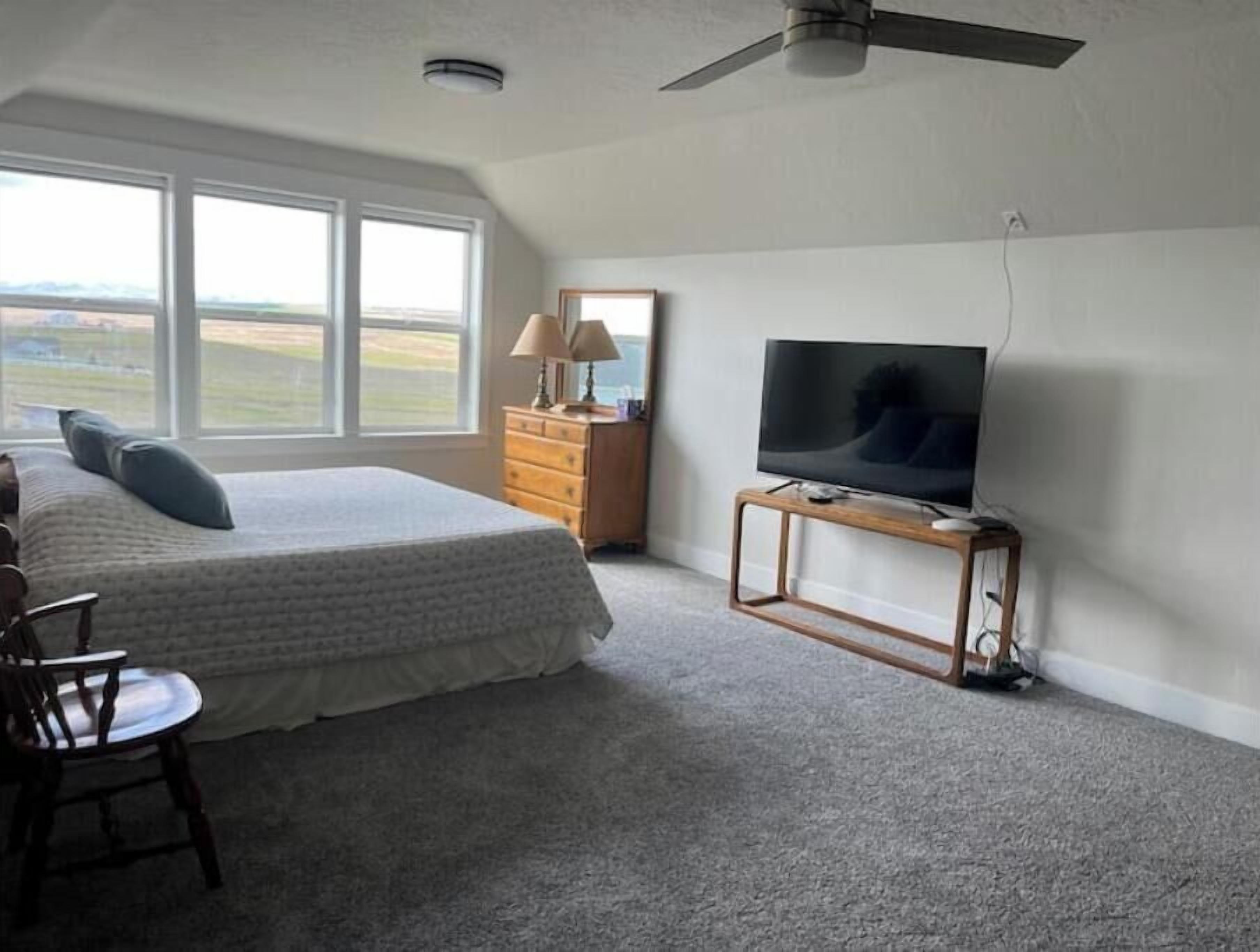 A bedroom with a large bed, a TV on a wooden stand, a dresser, and a view of the outdoors through large windows.