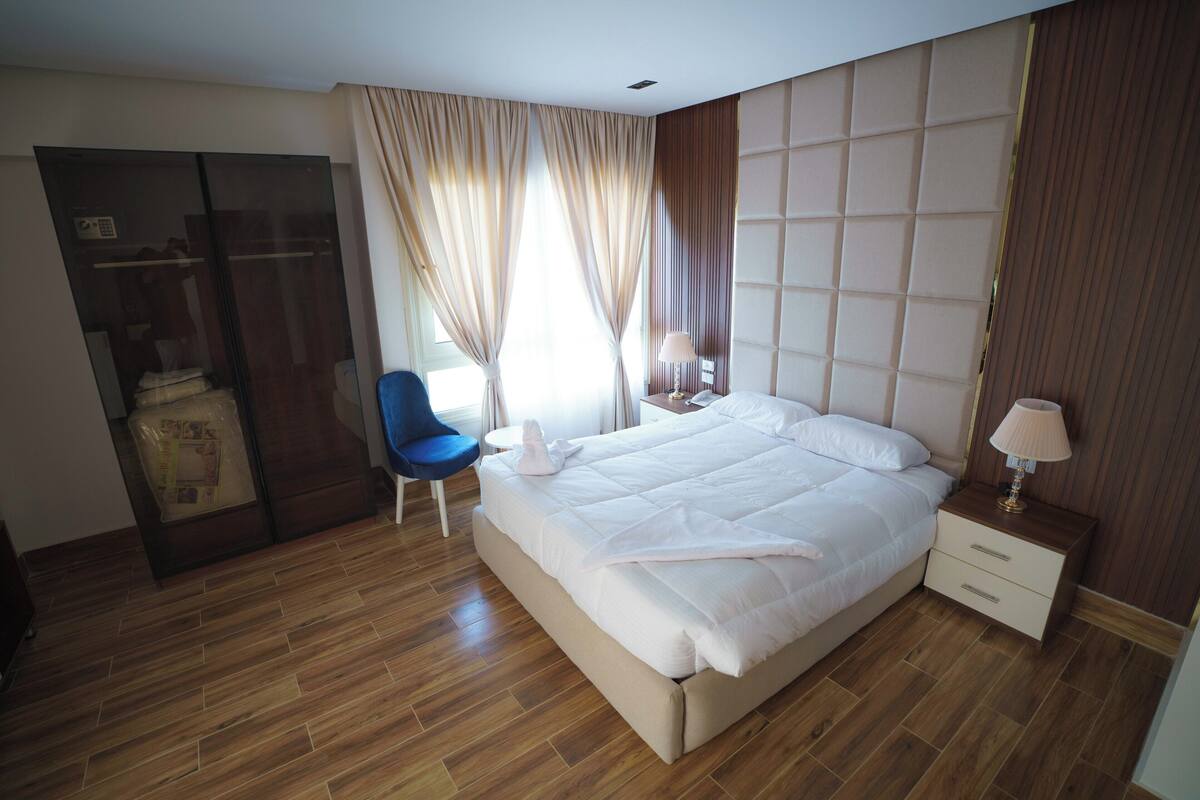 A modern hotel room with a large bed, a desk, a chair, and a wardrobe.