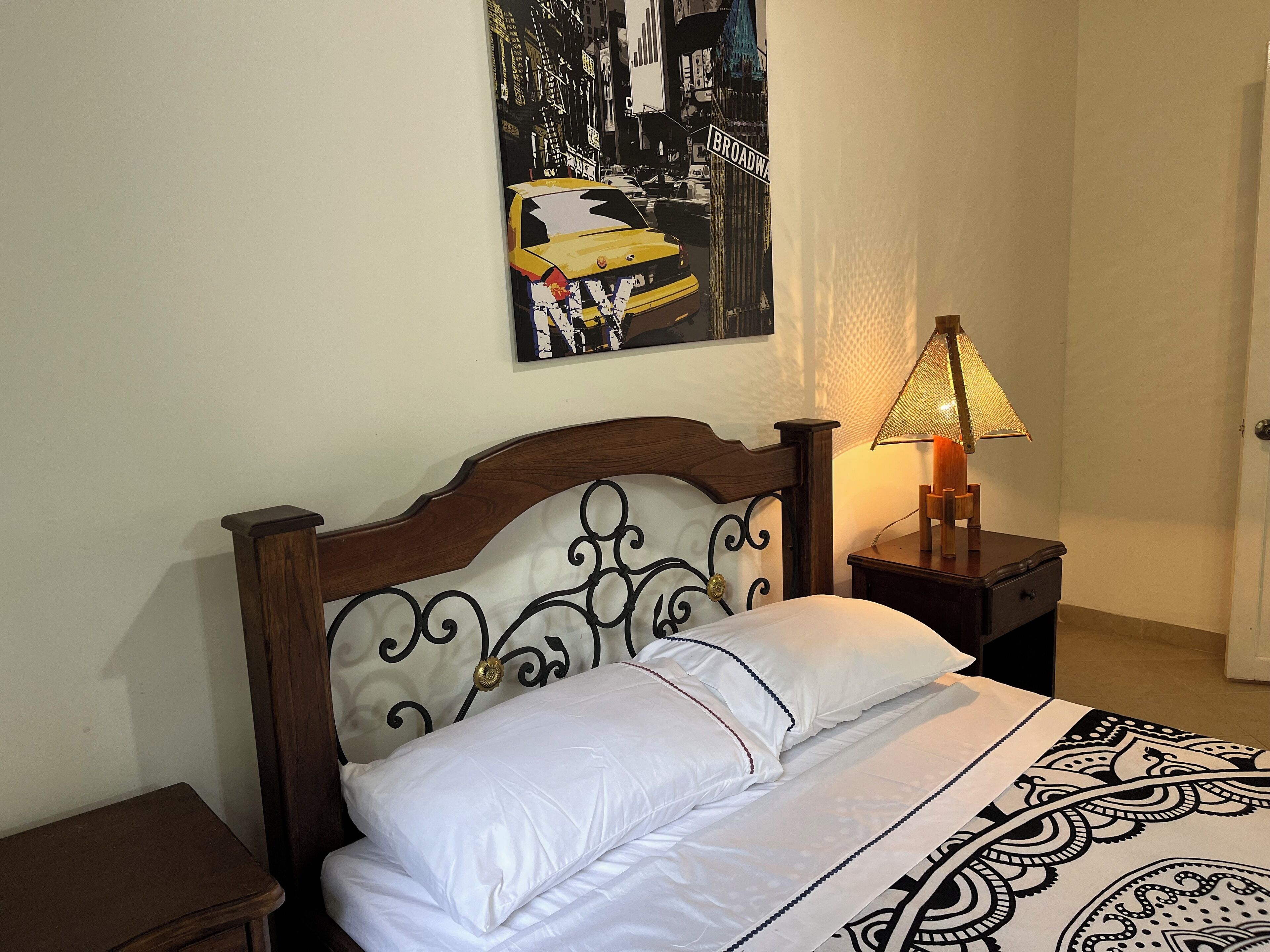 A neatly made bed with white linens, a decorative headboard, a wall-mounted artwork of a cityscape, and a bedside lamp.