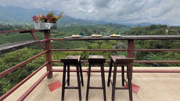 A wooden deck with a table and two chairs, overlooking a lush green landscape with mountains in the distance. - Finca Hotel San Miguel (Calarcá)