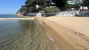 A beach with clear water, a sandy shore, and a building with a balcony overlooking the sea.