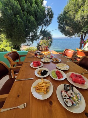 Una mesa de madera dispuesta con platos de comida, incluyendo sandía, aceitunas y queso, con vista a un cuerpo de agua.