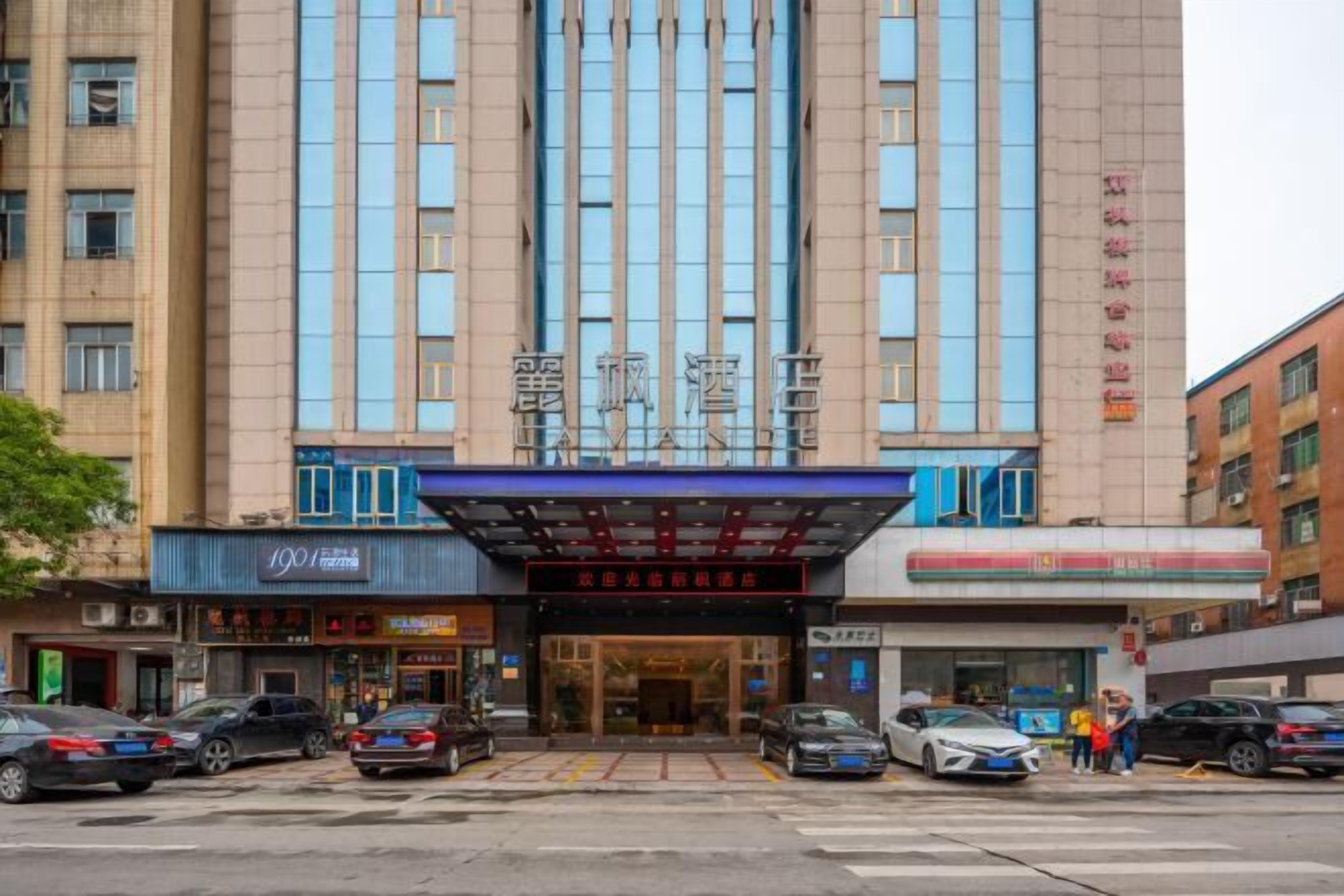 A multi-story hotel with a prominent entrance, several ground-level shops, and parked cars in front.