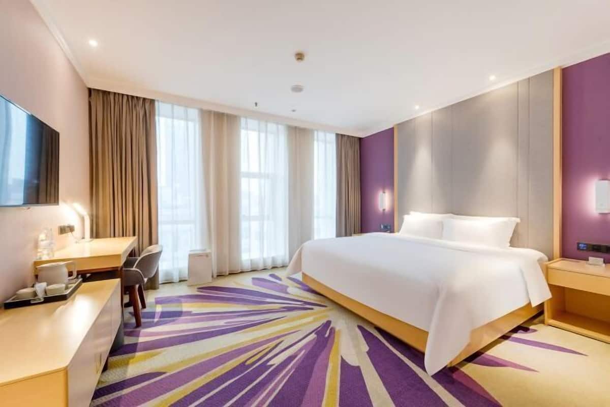 A hotel room with a large bed, a desk with a chair, a flat-screen TV, and a window with curtains.