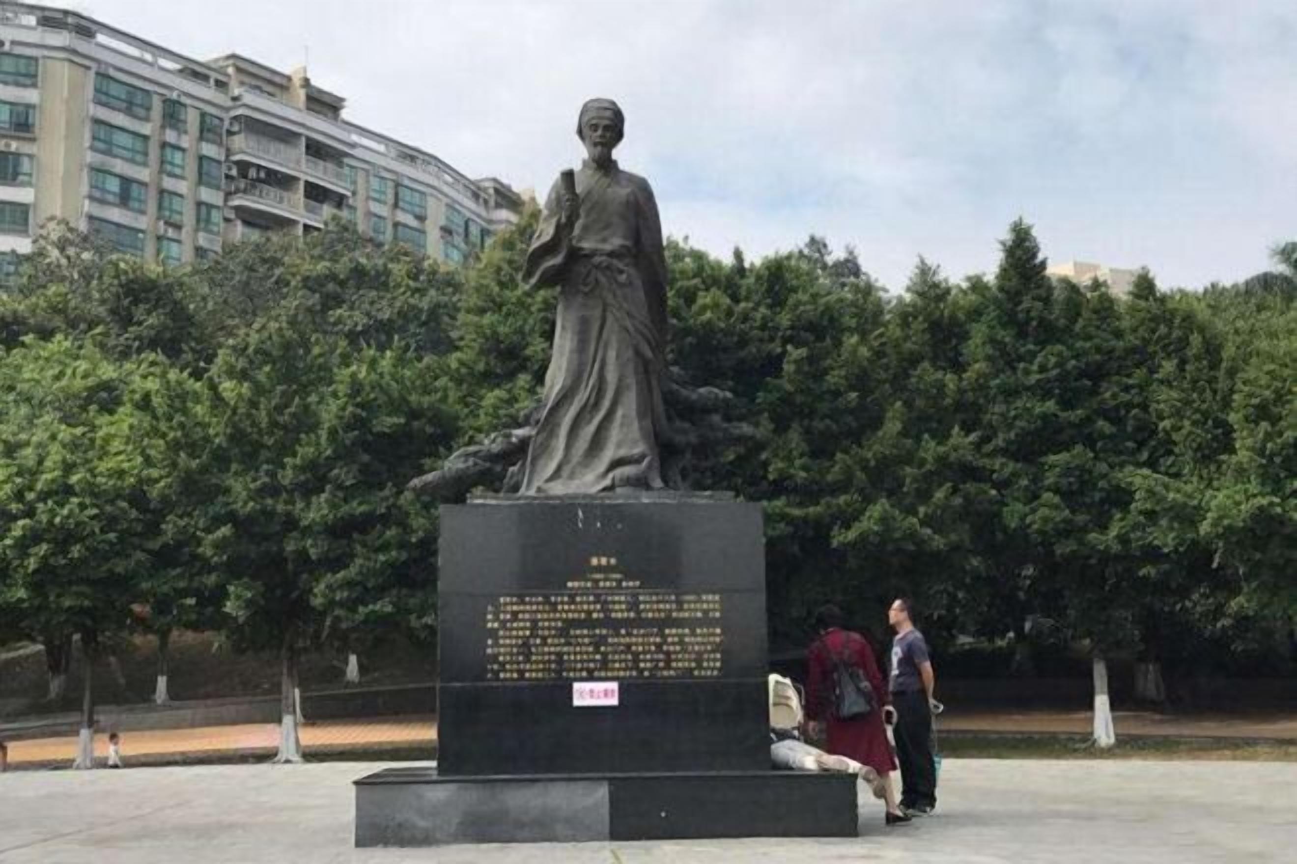 Una estatua al aire libre con una placa, rodeada de árboles y un edificio en el fondo.