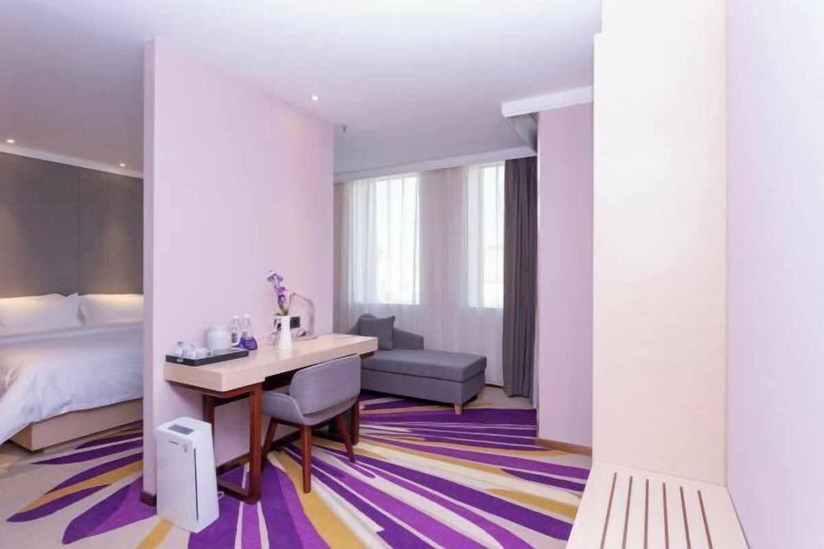 A hotel room with a bed, a desk with a chair, a sofa, and a large window with curtains.