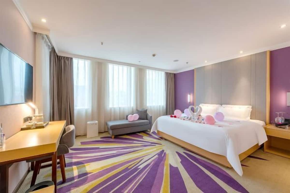 A hotel room with a bed, a desk, a chair, a sofa, and a large window with curtains.