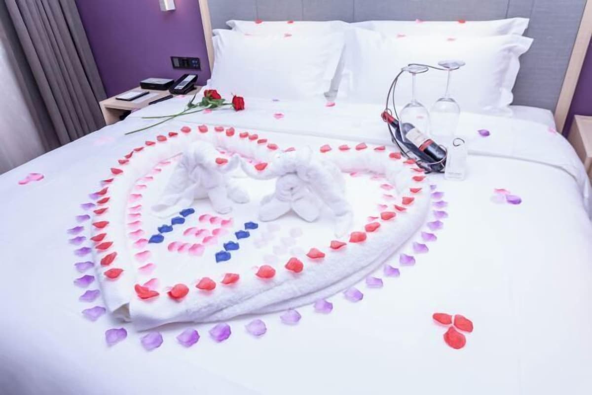 A hotel room with a neatly made bed, a bedside table with a phone and a book, and a decorative heart-shaped arrangement of rose petals and towels on the bed.