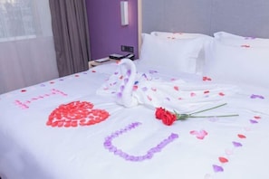 A neatly made bed with rose petals and a swan decoration, a purple wall, and a window with curtains.