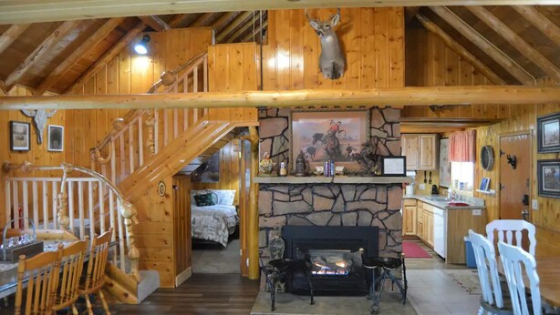 Een knus chaletinterieur met een stenen open haard, een houten eethoek en een trap naar een slaapkamer op een entresol.