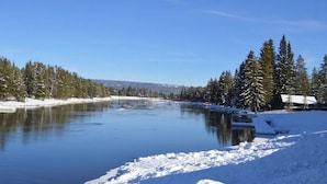 Een besneeuwd landschap met een bevroren rivier, dichte altijdgroene bomen en een hutje op de achtergrond.