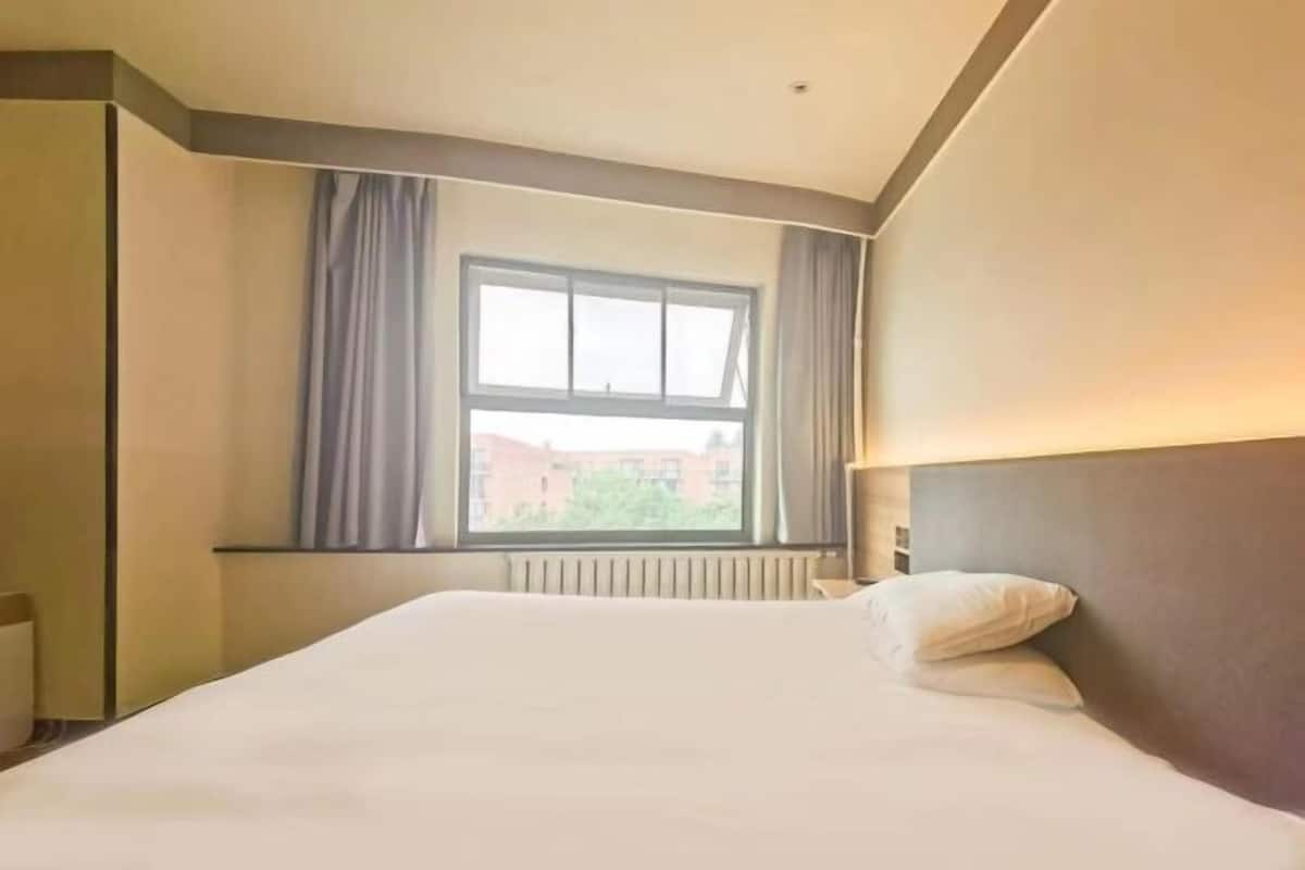 A hotel room with a large bed, a window with curtains, and a view of buildings outside.