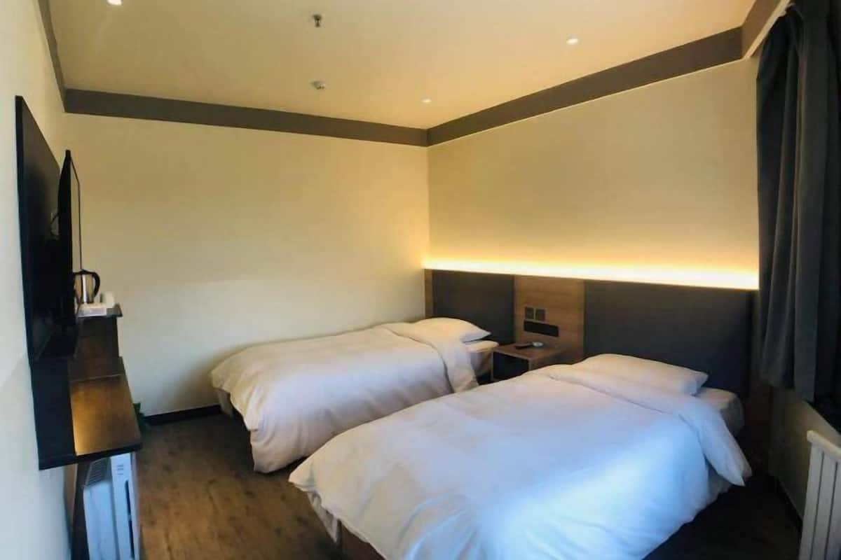 A hotel room with two beds, a desk, a flat-screen TV, and a wall-mounted light.