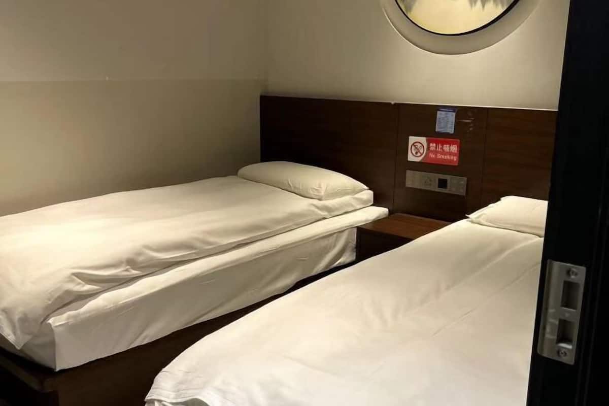 A small, private room with two single beds, a wooden headboard, a round mirror, and a no-smoking sign.
