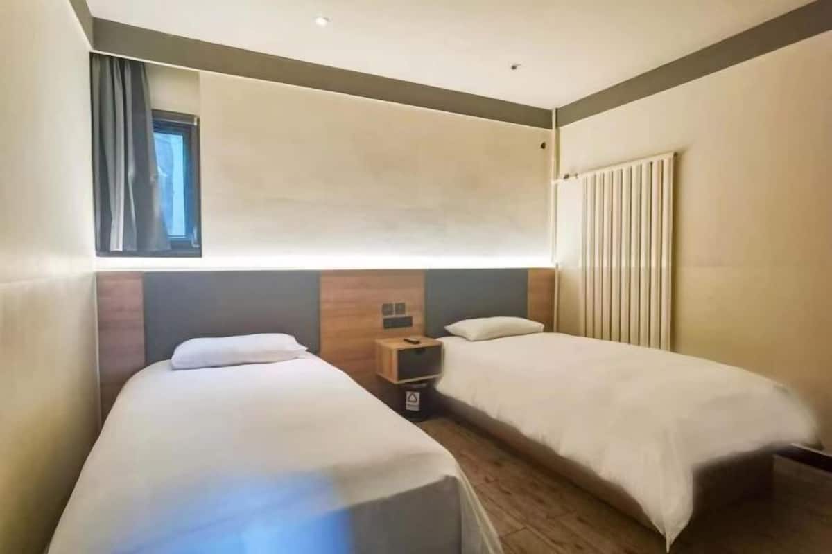 A hotel room with two single beds, a wooden headboard, a nightstand with a lamp, and a window with curtains.