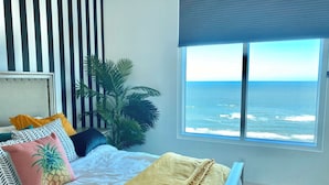 Een slaapkamer met een bed, een groot raam met uitzicht op de oceaan, een kamerplant en gestreepte wanddecoratie.