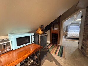 A compact kitchenette with a microwave, toaster, and small refrigerator. A wooden cabinet with a knife block is visible, and a bed is seen in the adjacent room.